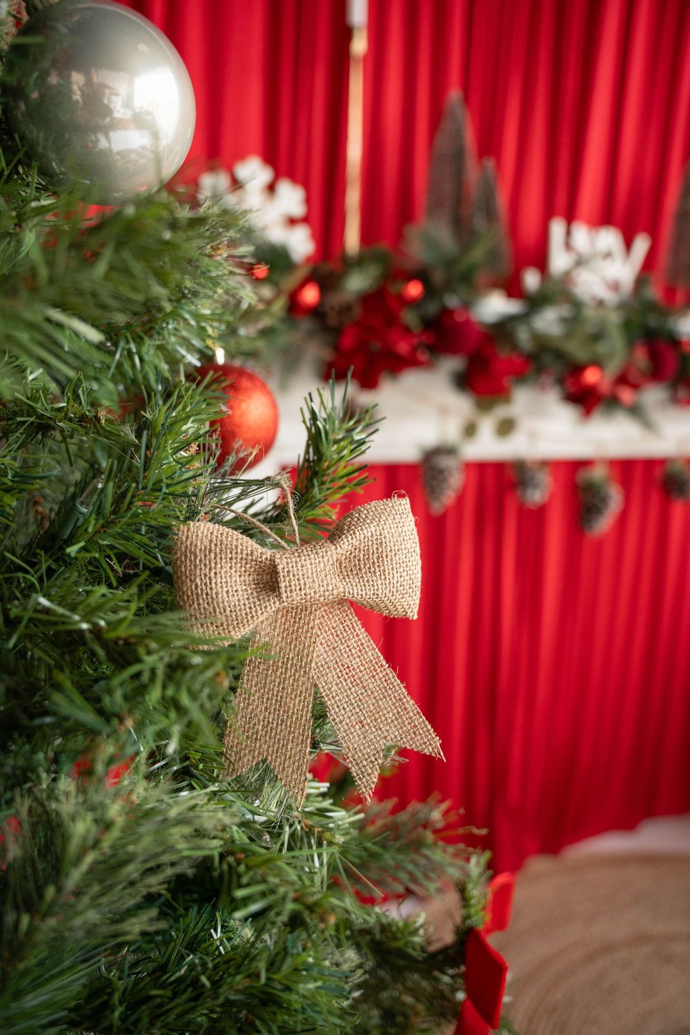 Close-up of a Christmas tree with burlap bow, red and silver ornaments, and festive garland against red curtains.