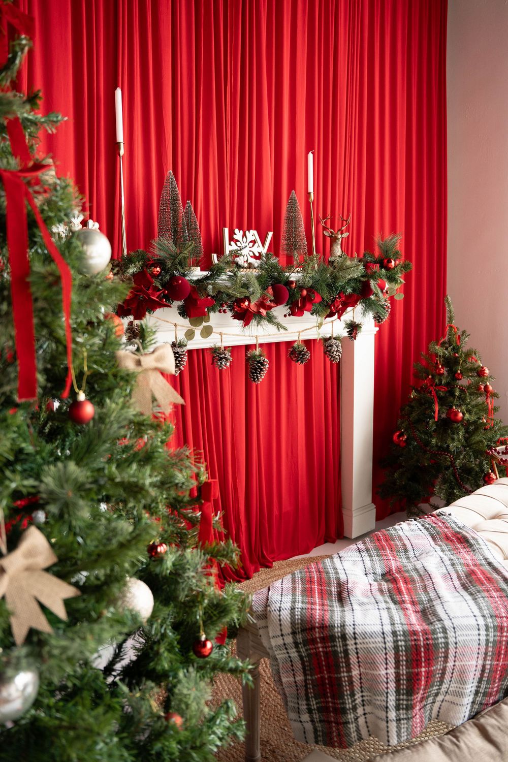 Festive Christmas room with decorated fireplace mantel, Christmas trees, red curtains, candles, and plaid ottoman.