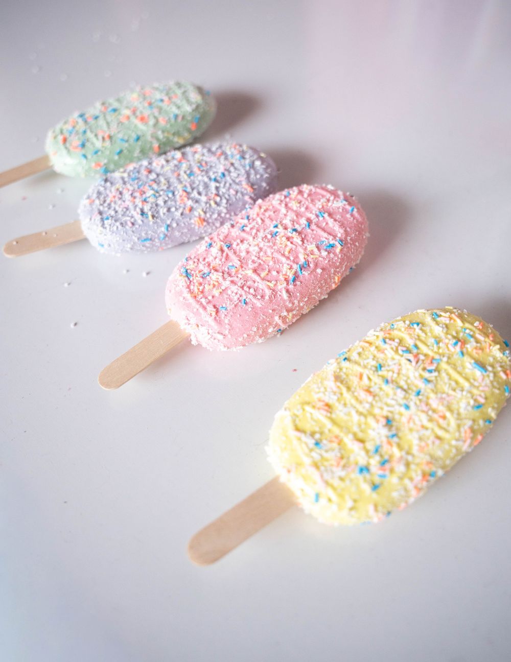 Four pastel-colored ice cream bars with rainbow sprinkles on wooden sticks arranged on a white surface.