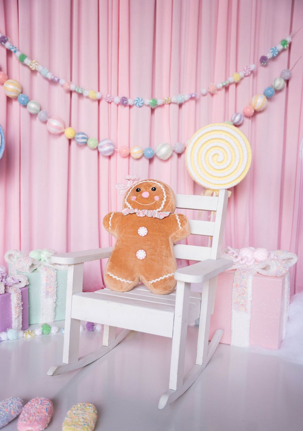 Pastel pink candy-themed backdrop with gingerbread man plush on white rocking chair, lollipop decor, and colorful garland.