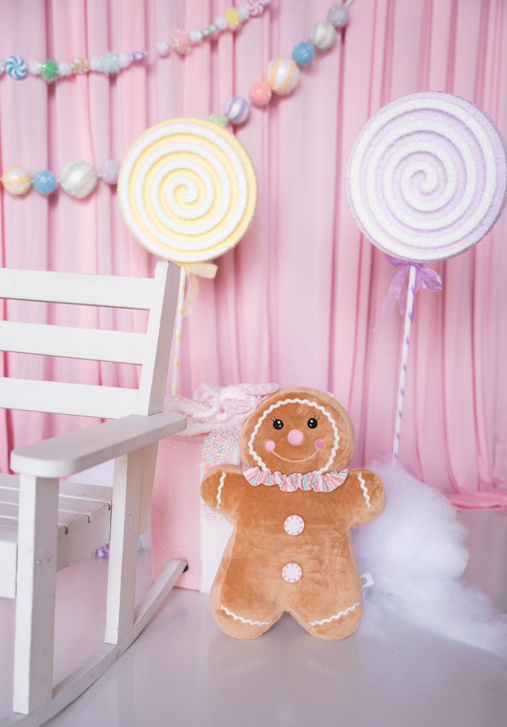 Candy-themed photo backdrop with gingerbread man plush, lollipop props, pastel pink curtains, and white chair.