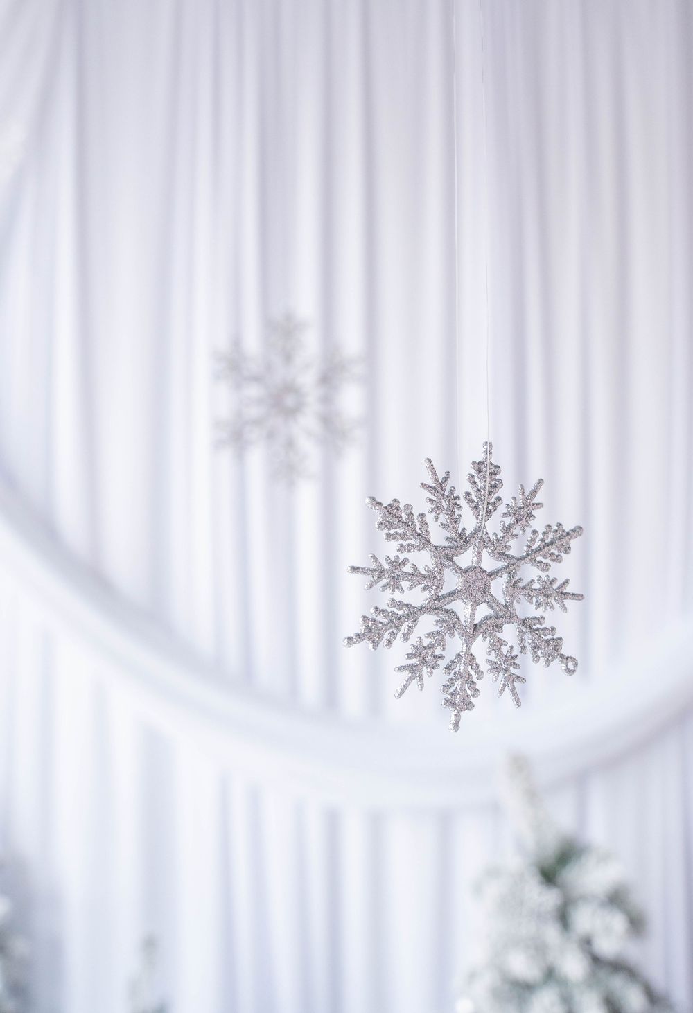 Silver glitter snowflake ornaments hanging against white draped fabric backdrop with Christmas tree decoration.