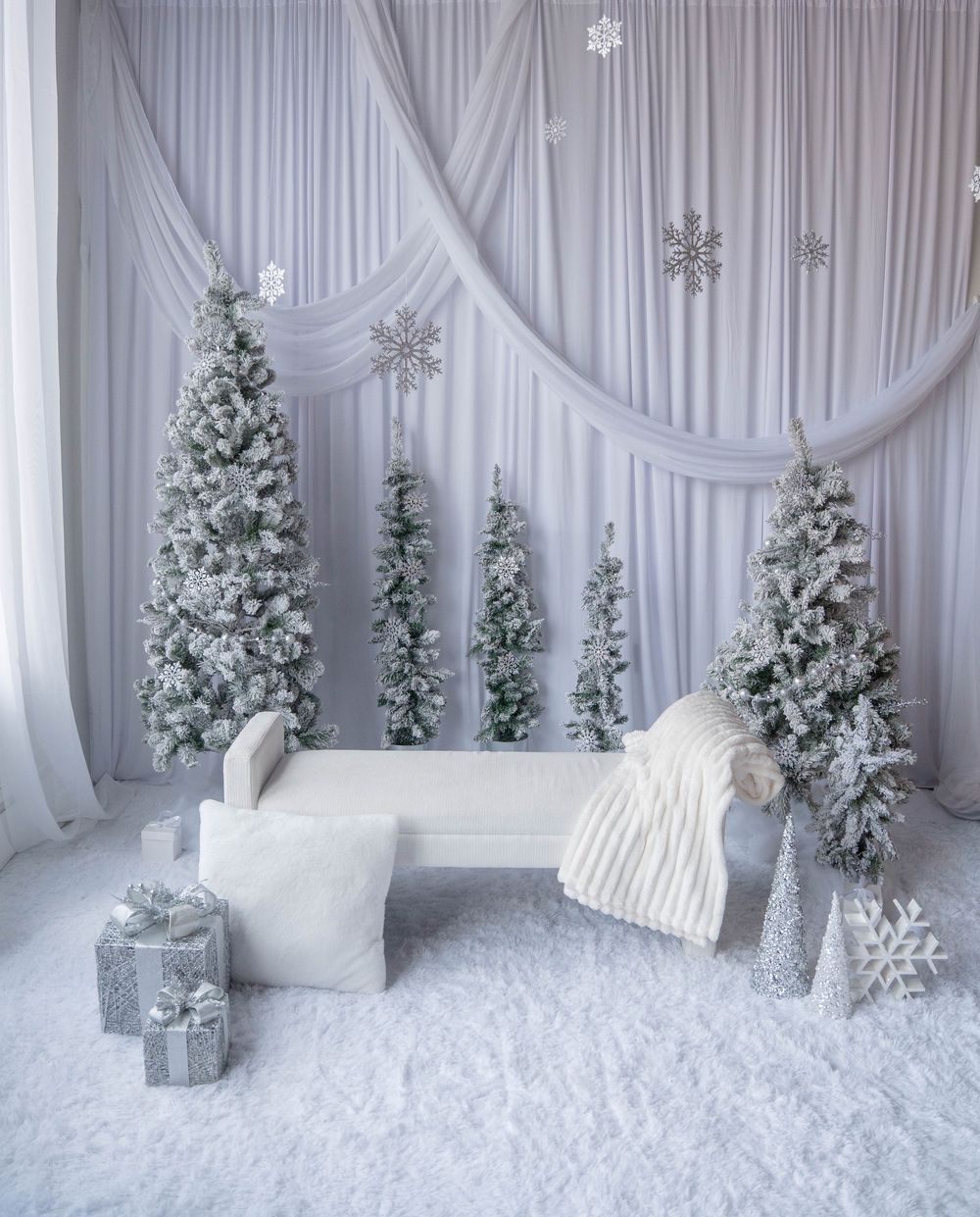 White winter wonderland Christmas scene with flocked trees, silver gifts, snowflakes, and cozy bench with knit throw.