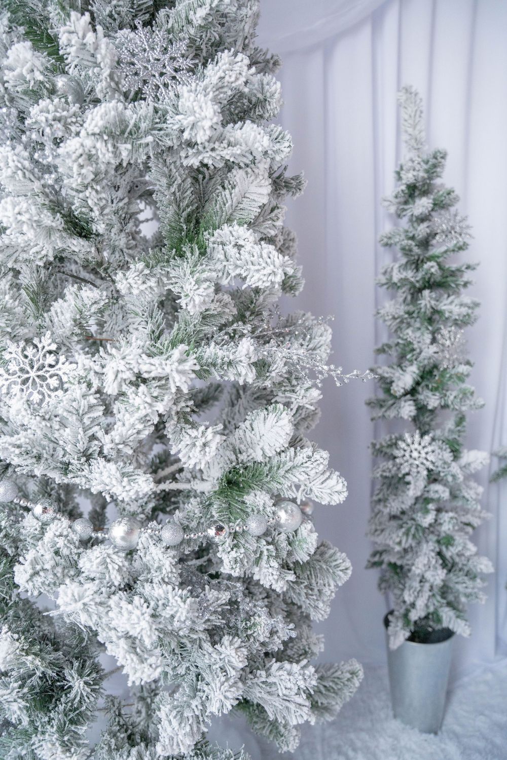 Two snow-flocked artificial Christmas trees decorated with white snowflake ornaments in a bright white holiday room setting.