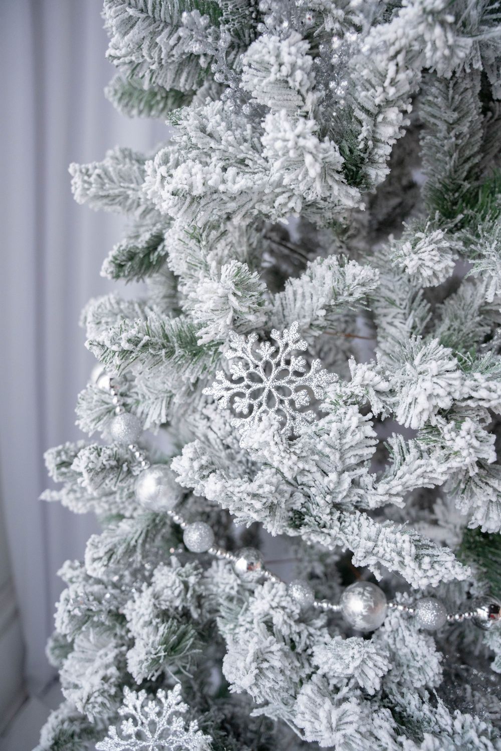 Snow-flocked Christmas tree decorated with silver snowflake ornaments and baubles in elegant white winter holiday decor.