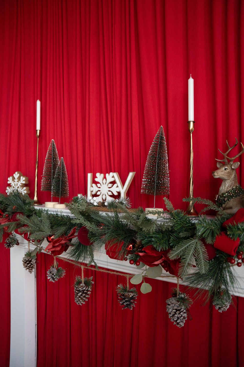Christmas mantel with pine garland, pinecones, candles, mini trees, JOY sign, and reindeer figurine against red curtain backdrop.