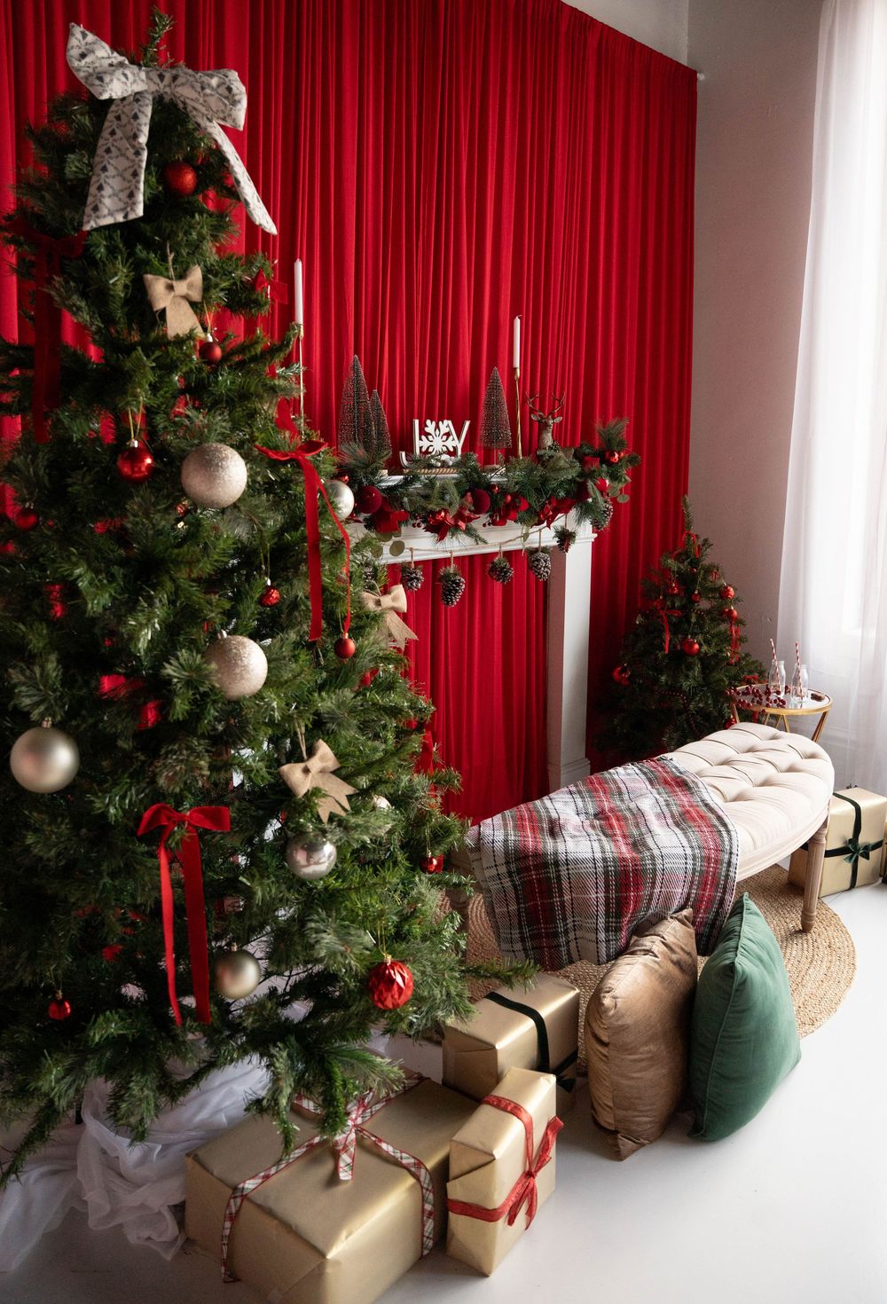 Festive Christmas room with decorated tree, wrapped gifts, plaid bench, and red curtain backdrop with mantel display.