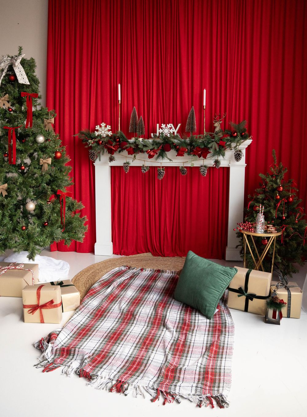 Festive Christmas scene with red curtains, decorated fireplace mantel, plaid blanket, green pillow, wrapped gifts, and lit Christmas tree.