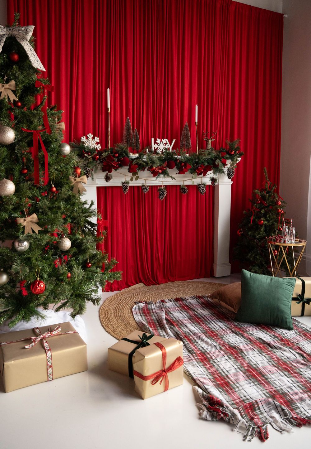 Festive Christmas living room with decorated tree, wrapped gifts, plaid blanket, and red curtain backdrop with garland mantle.