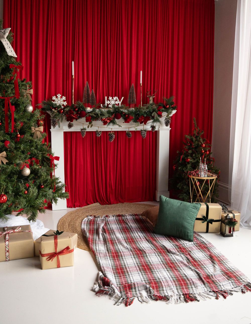 Festive Christmas room with red curtains, decorated fireplace mantel, plaid blanket, green cushion, and wrapped gifts under Christmas trees.