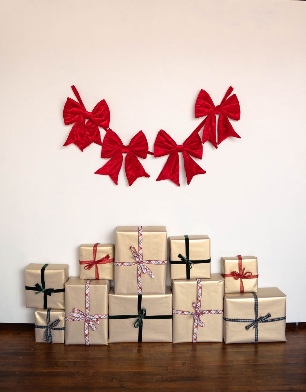Stack of kraft paper wrapped Christmas gifts with colorful ribbons beneath red velvet bows garland on white wall.