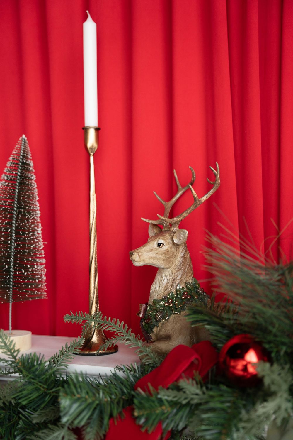 Christmas deer figurine with gold candlestick, mini tree, and red ornaments against red curtain backdrop.
