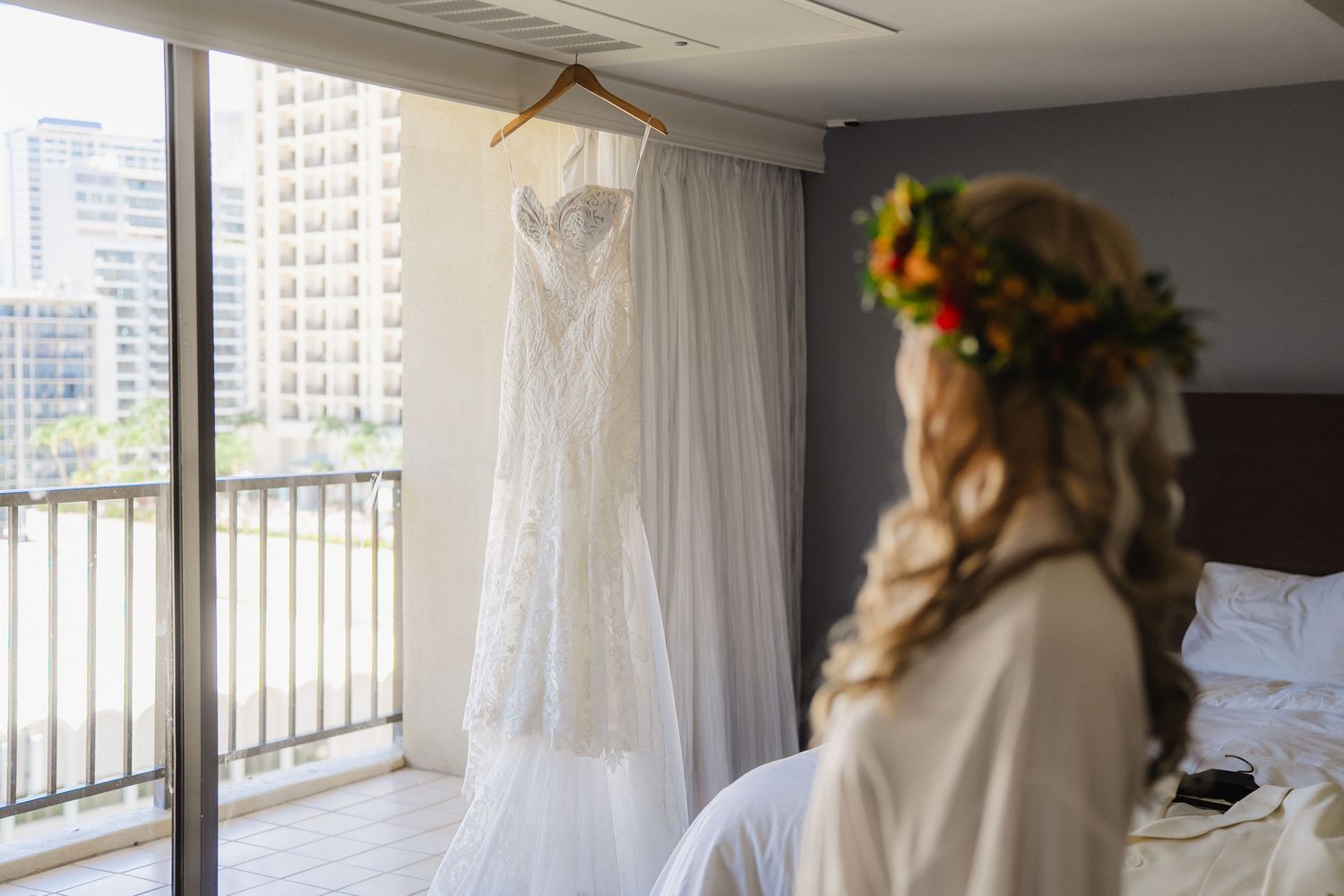 Bride in floral crown views her white lace wedding dress hanging by a balcony window in a hotel room.