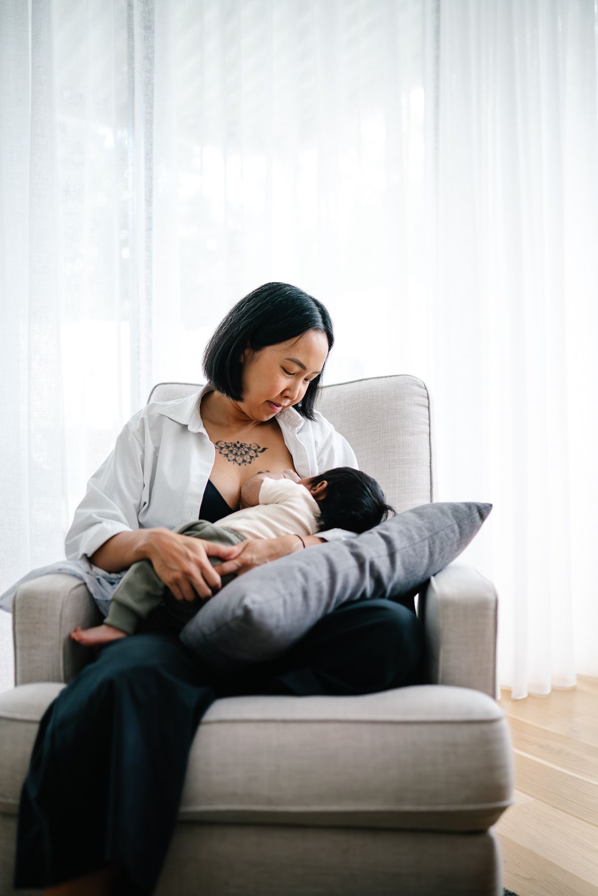 A mother sits in a cozy armchair breastfeeding her infant, in a bright, serene room taken by a Melbourne in-home Newborn photographer.