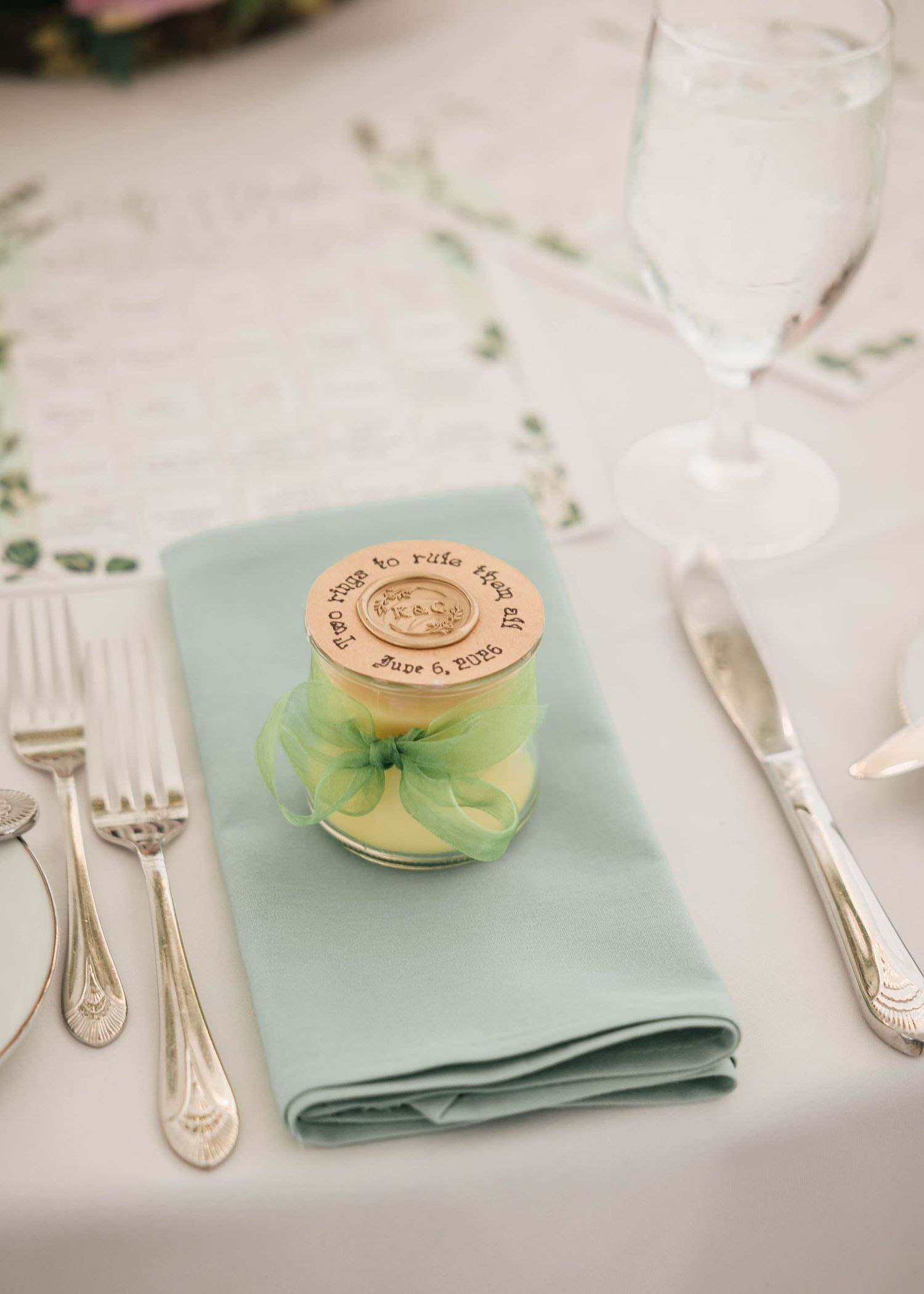 Wedding favor with a wax-sealed tag reading 'two rings to rule them all,' tied with green ribbon on a place setting