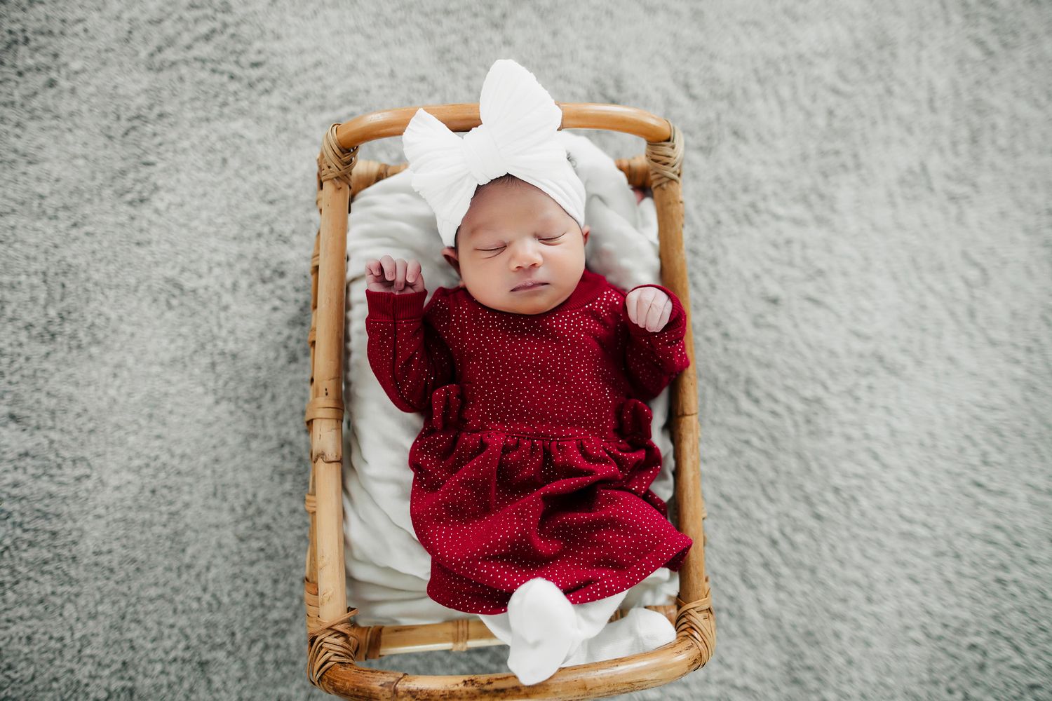 Newborn baby sleeping in wicker basket wearing red knit outfit and white bow headband on gray background.