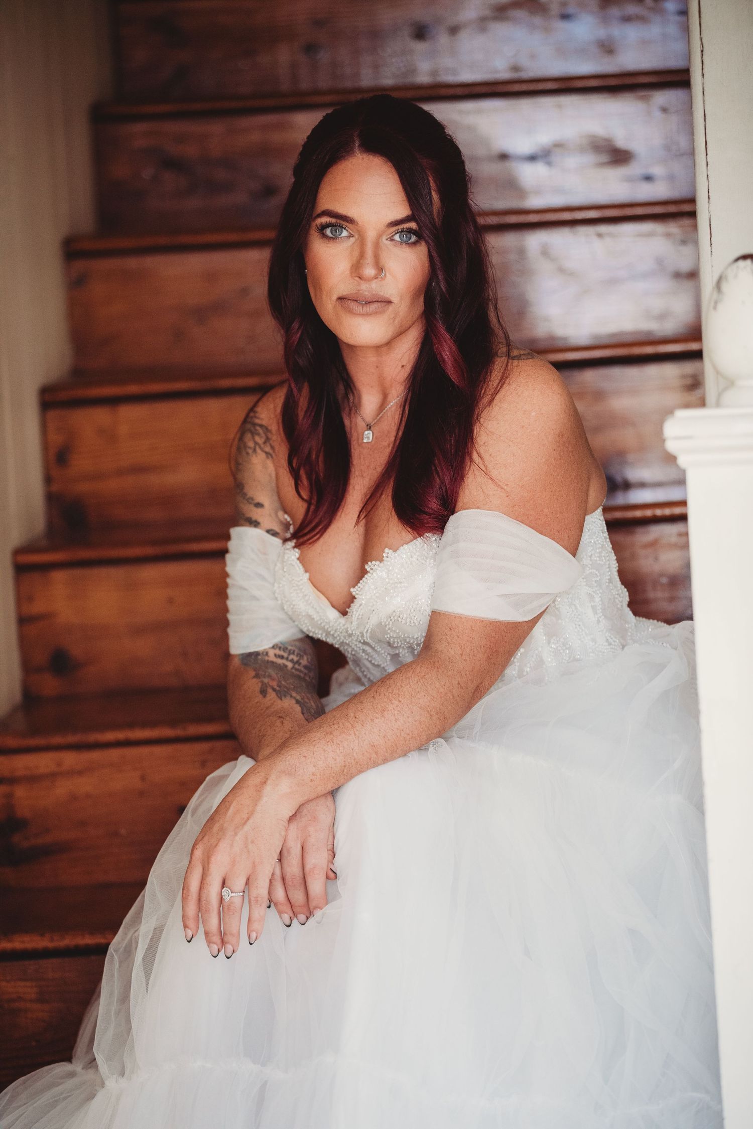 A tattooed bride in an off-shoulder white tulle gown sits gracefully on wooden stairs, wearing a diamond ring.