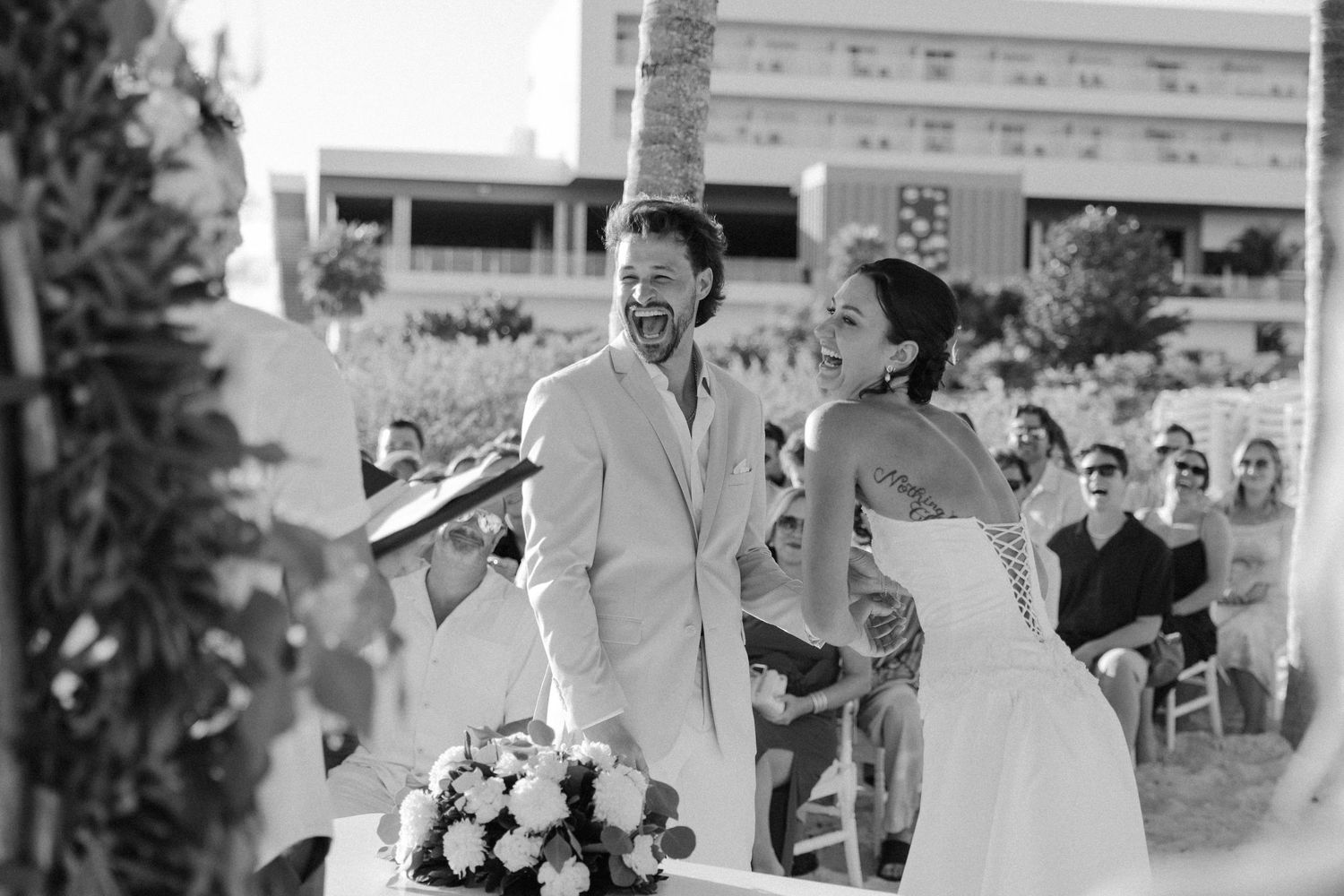 Joyful bride and groom laughing during outdoor wedding ceremony with guests seated in background.