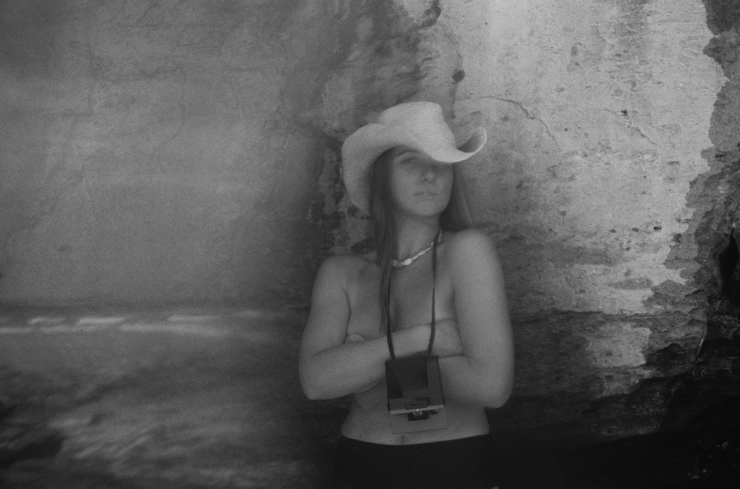 Person wearing a cowboy hat eating a banana while swimming in calm ocean waters near a sandy beach.