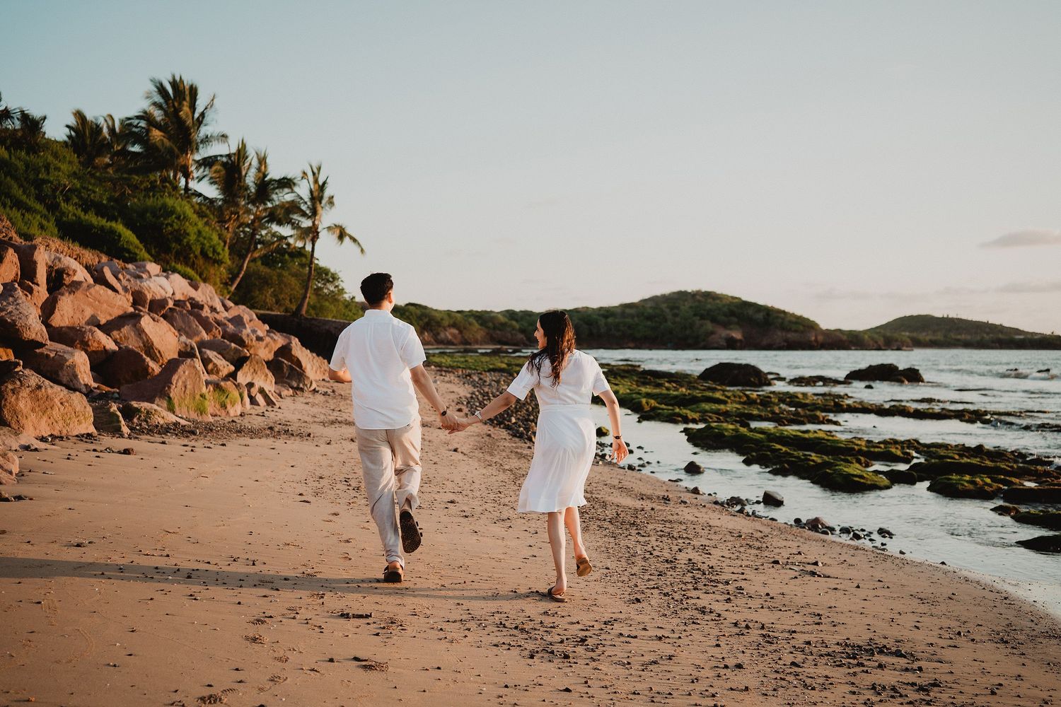 Pareja caminando entre rocas durante una sesión de fotos en la playa.