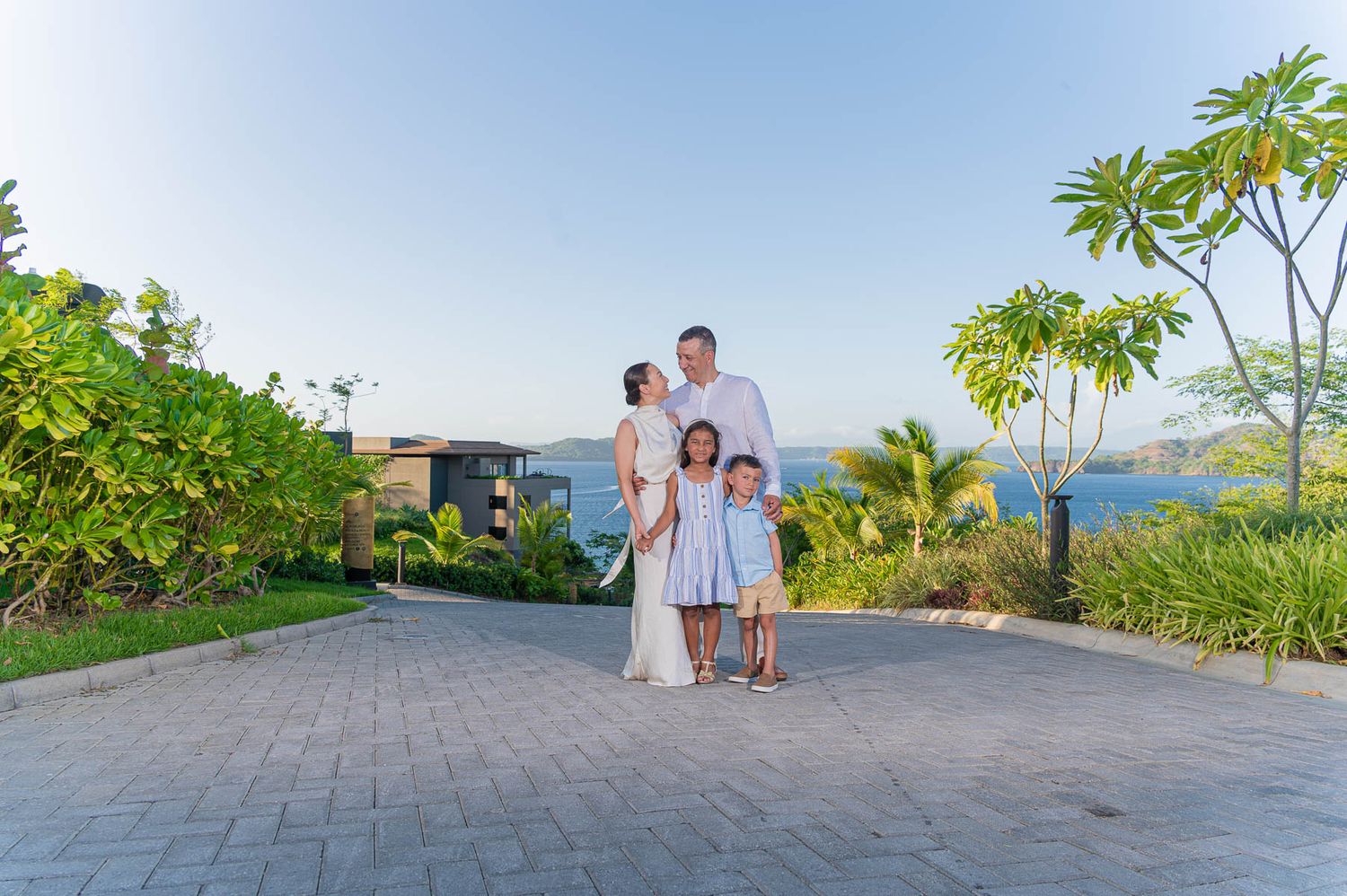 Family of four posing on a paved path at a tropical resort with lush greenery and ocean views at sunset.