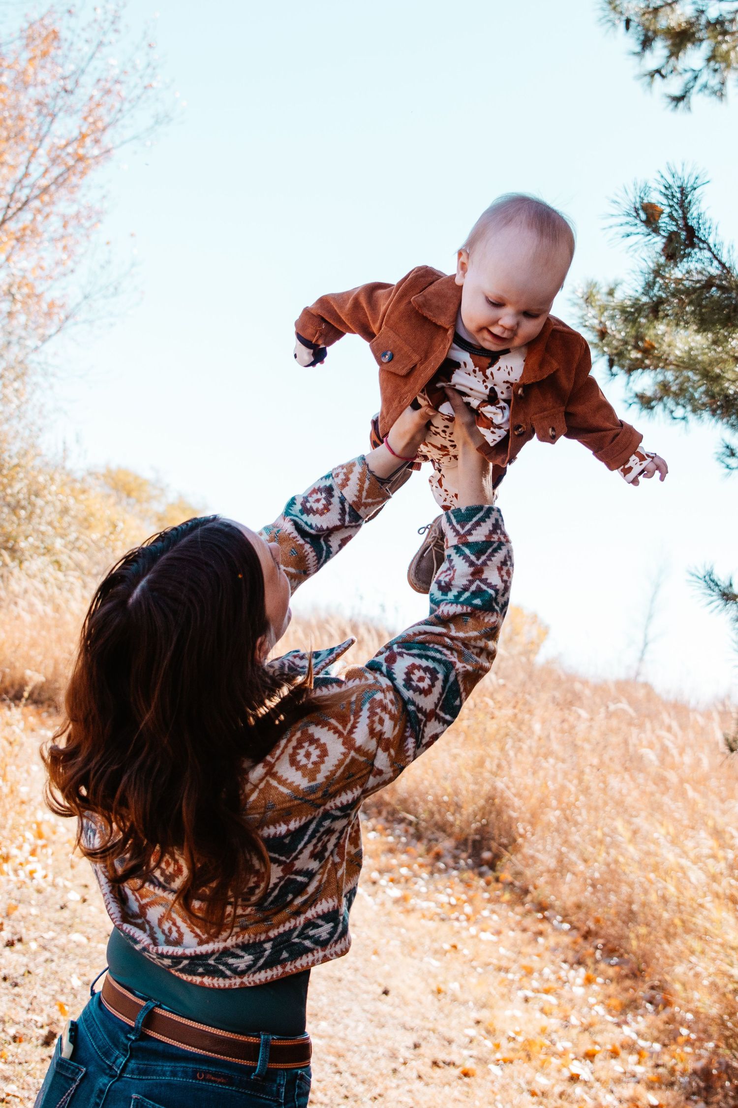 A mother joyfully lifts her baby in the air outdoors during autumn, both wearing matching boho-style outfits.