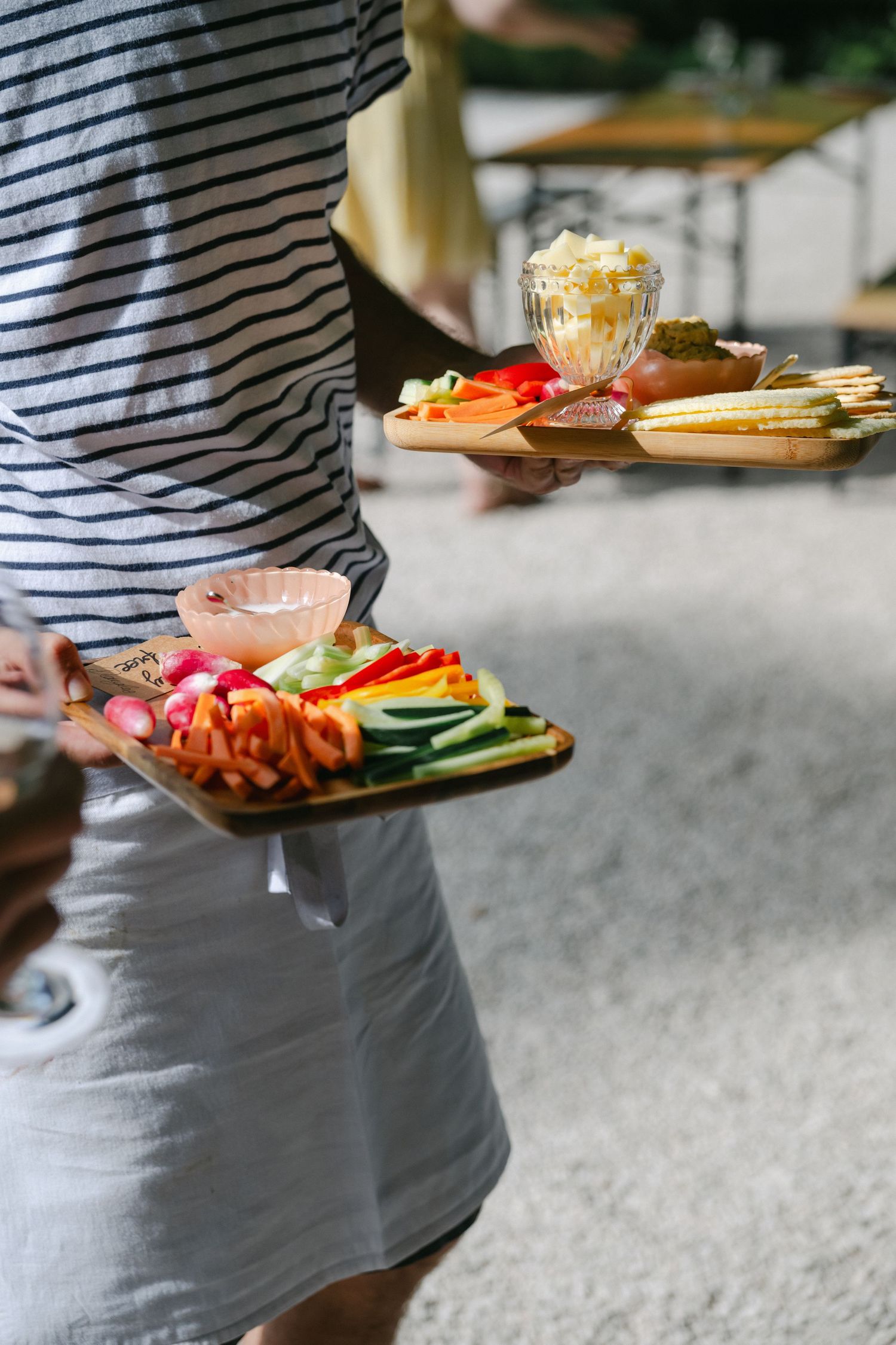 Person carrying two wooden boards with colorful sliced vegetables and dips at an outdoor party gathering.