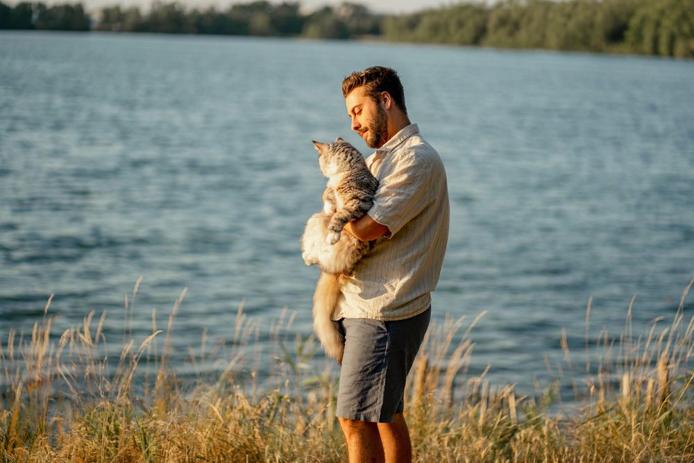 Un homme souriant tenant son chat au bord d'un lac au coucher de soleil