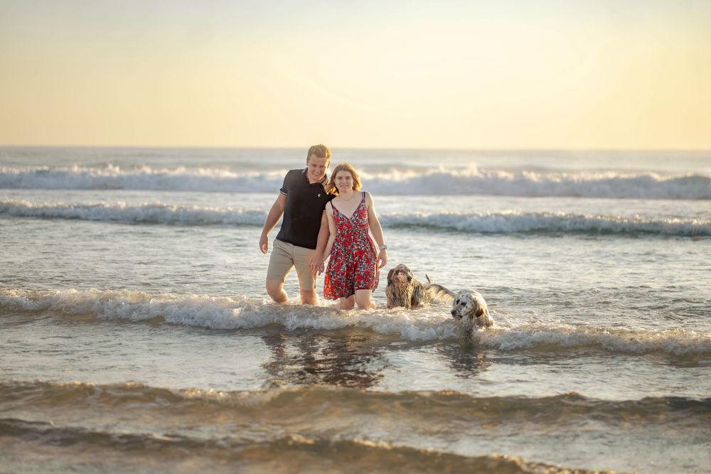 Un couple marchent dans les vagues de l'océan au coucher du soleil avec leur chien