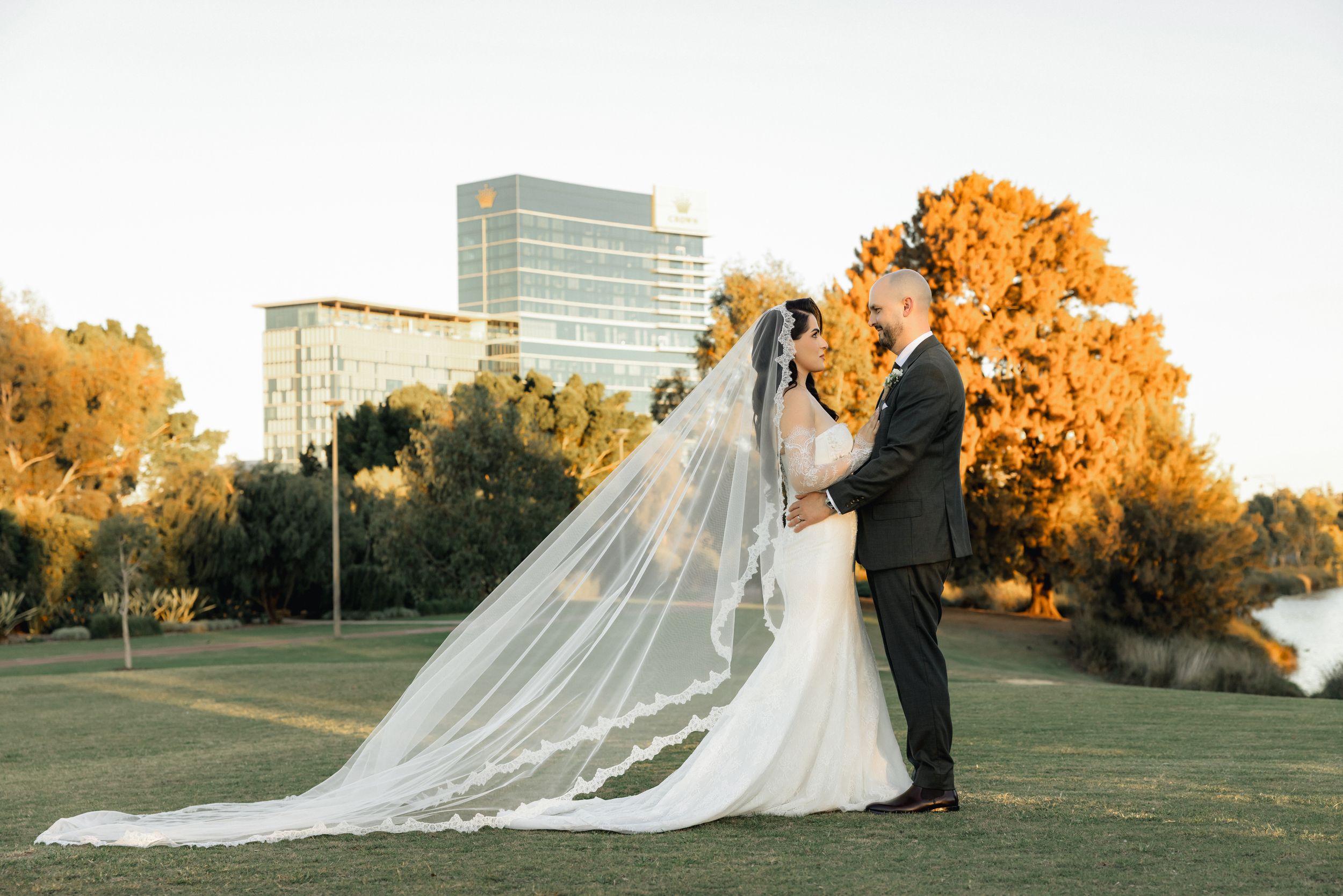 Bride and groom share a romantic kiss on a lush green lawn, with a flowing cathedral veil and golden sunset trees.