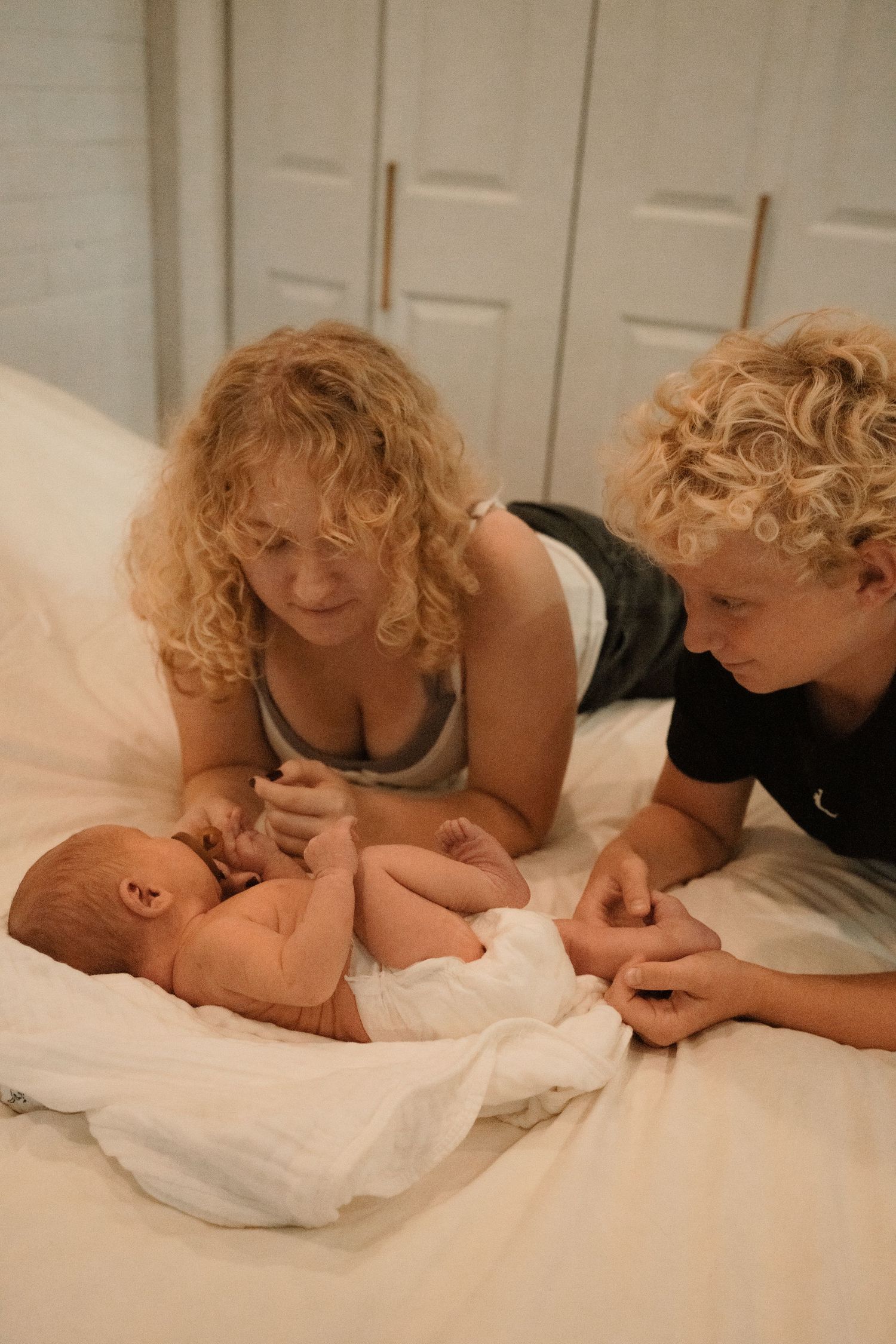 A woman and curly-haired child lying on a white bed, lovingly gazing at a newborn baby wrapped in a white blanket.