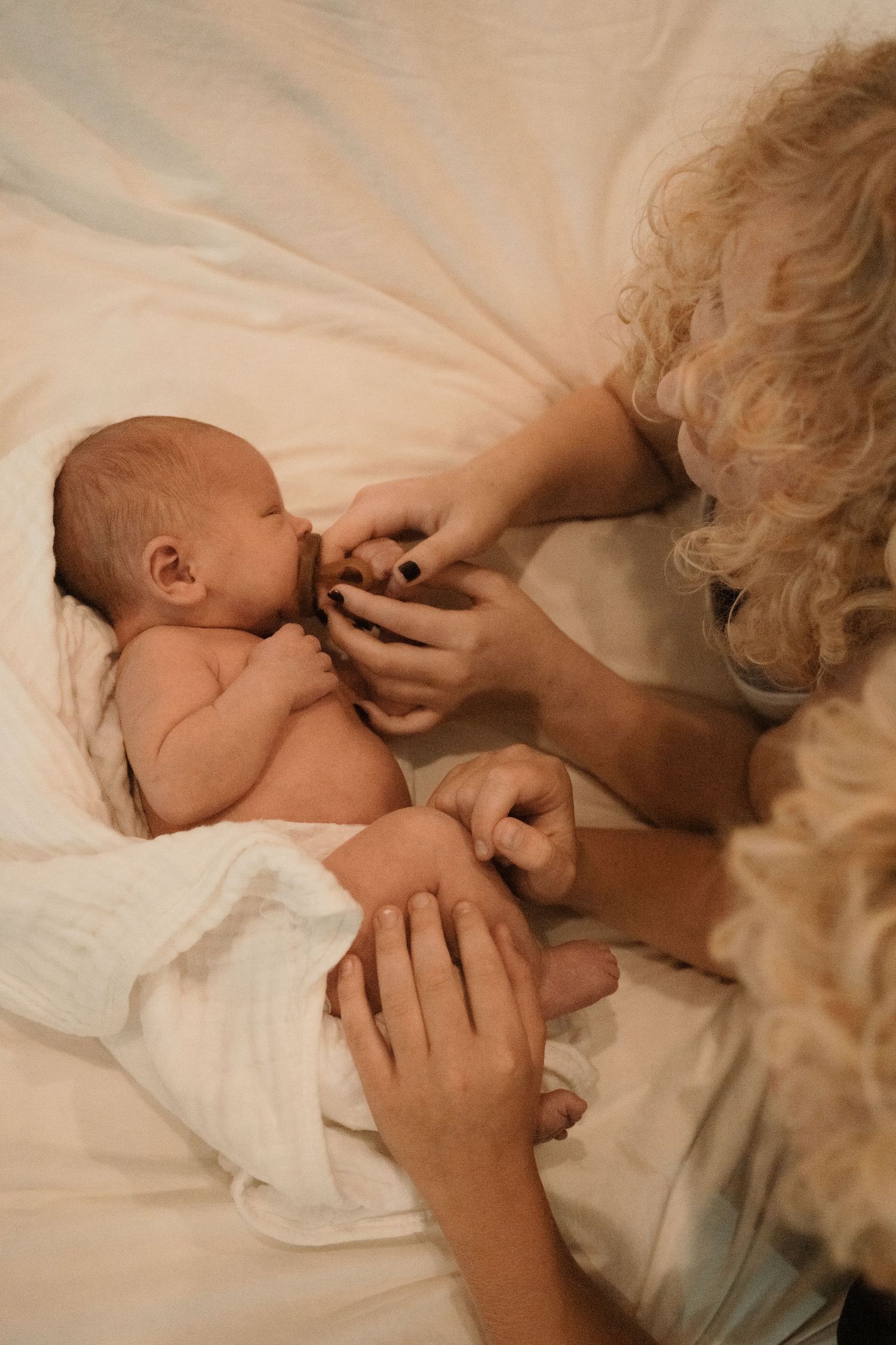 Curly-haired woman tenderly caring for a newborn baby lying on white bedding.