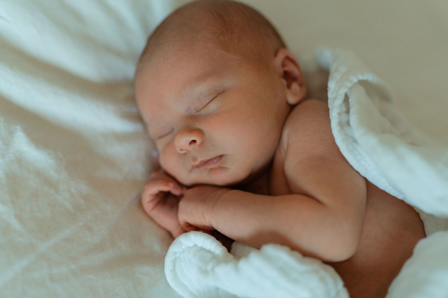 Peaceful newborn baby sleeping soundly on white blanket, eyes closed, tiny hands tucked under chin.