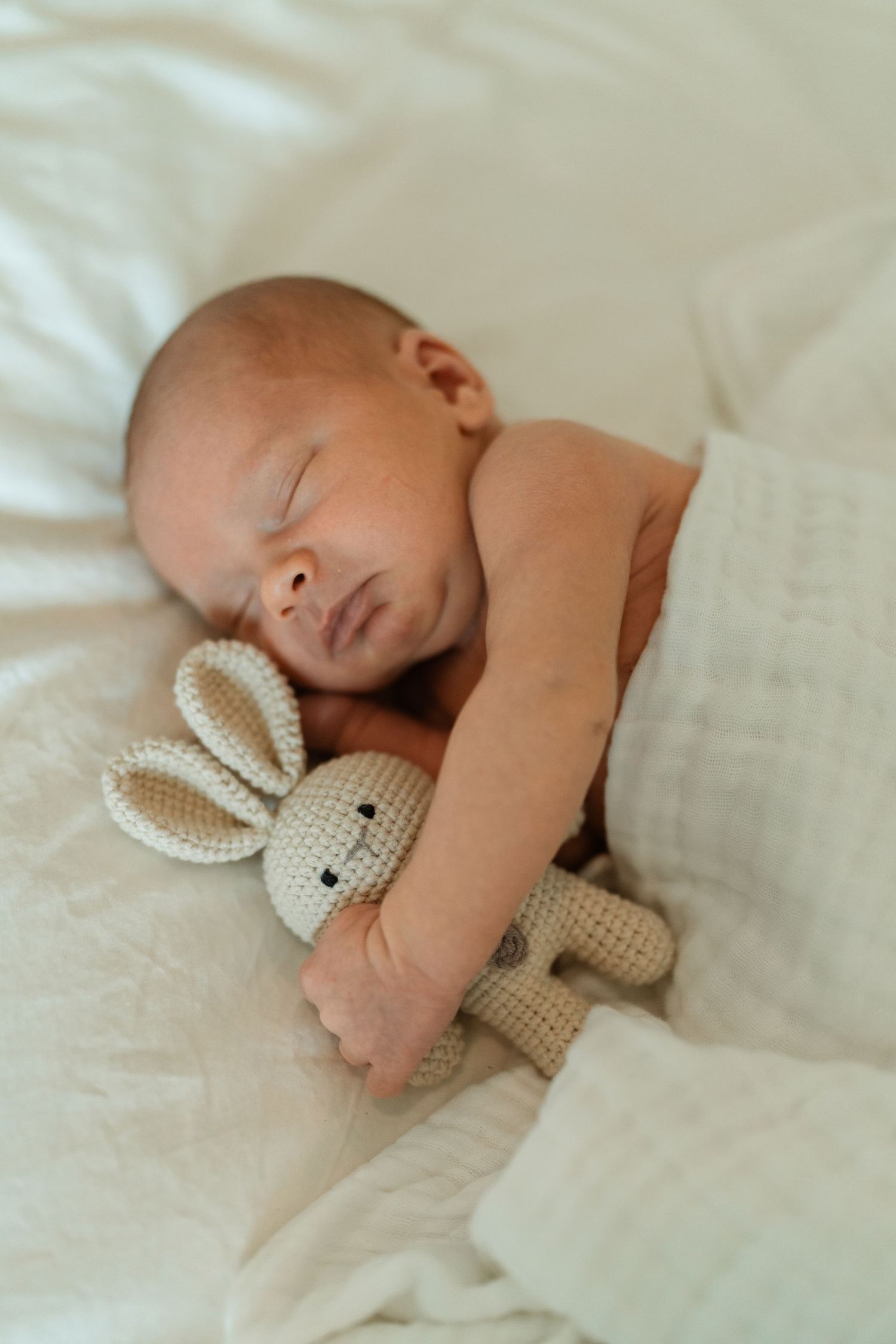 Newborn baby sleeping peacefully on white bedding while cuddling a small crocheted bunny stuffed animal toy.