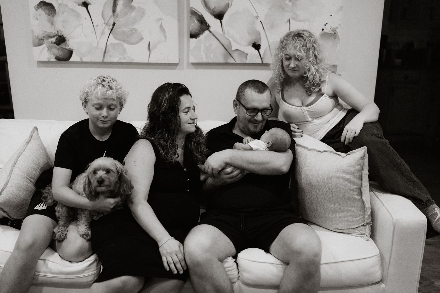 Black and white family photo of four adults and a dog sitting on a white sofa, with a newborn baby being held.