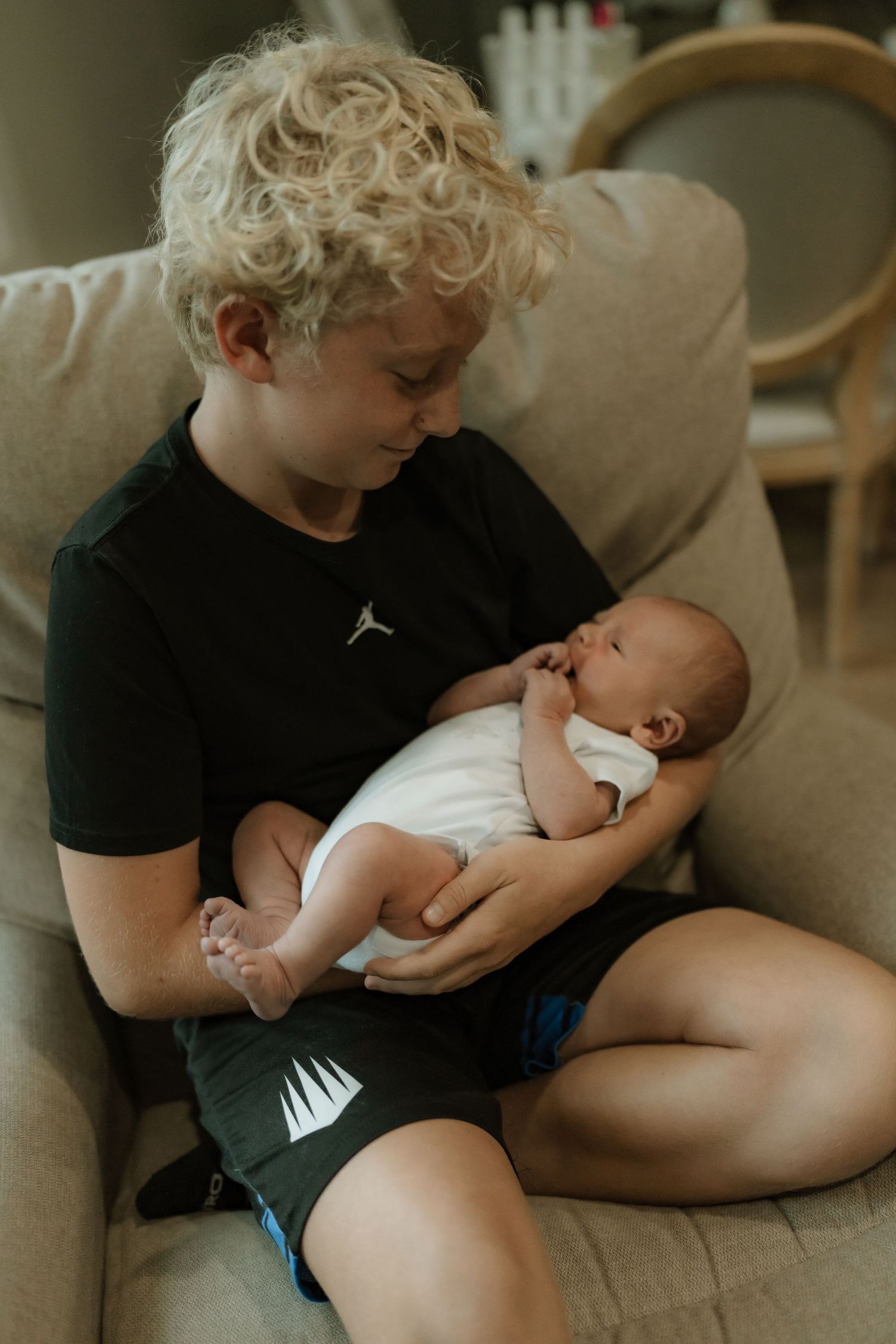 A young blonde curly-haired boy sits in a chair carefully holding a newborn baby in his arms.