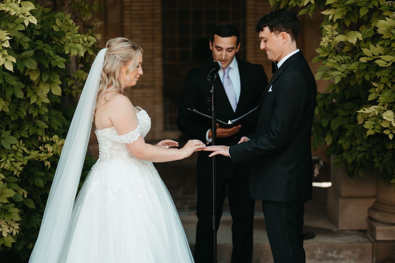 Bride and groom exchanging rings during outdoor wedding ceremony with officiant, surrounded by lush green ivy backdrop.