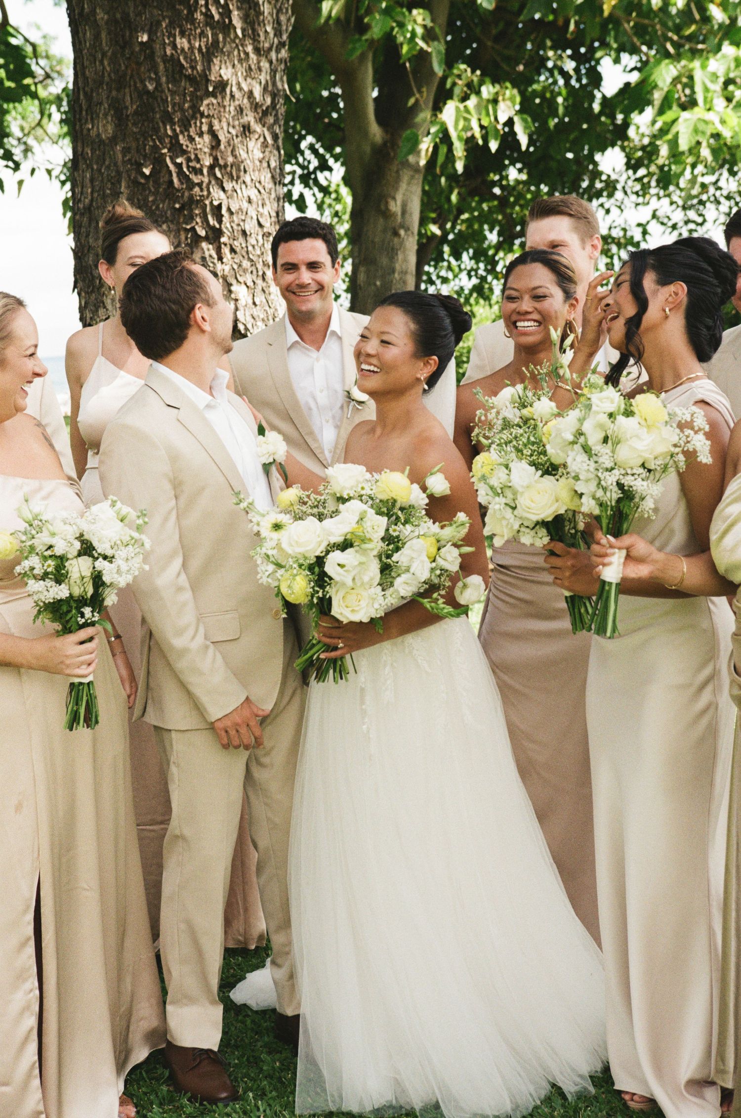 Wedding party in champagne attire laughing outdoors, bride in white tulle gown holding white floral bouquet with bridesmaids.