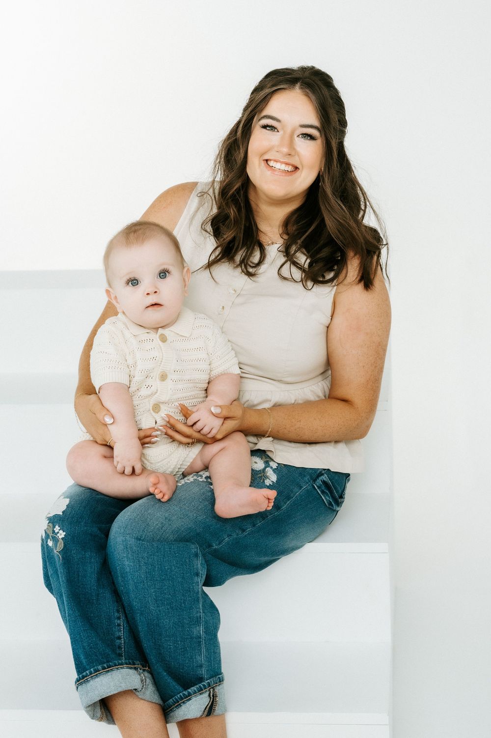 A smiling young woman with dark hair sits on white steps holding a cute baby in a knit outfit, both dressed casually.