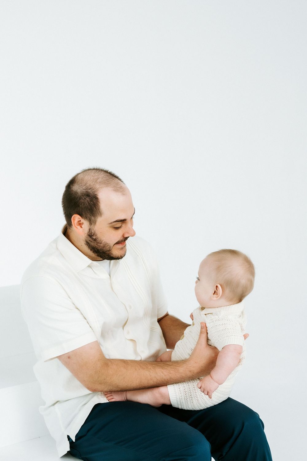A bearded father in a white shirt holds his baby infant while sitting against a white background.