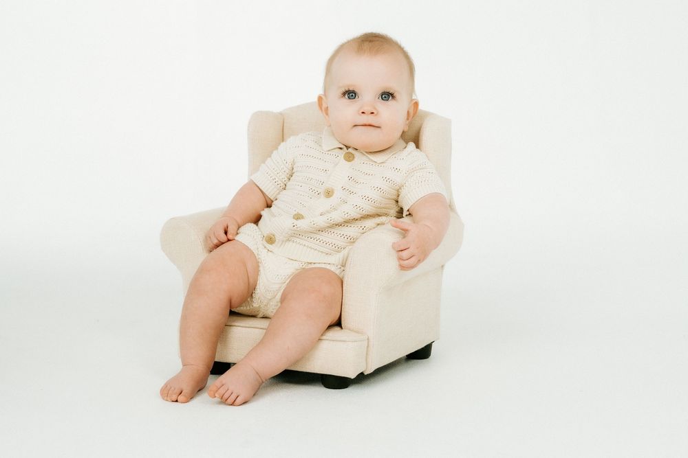Adorable baby sitting in a small cream armchair, wearing a white romper, against a white background.