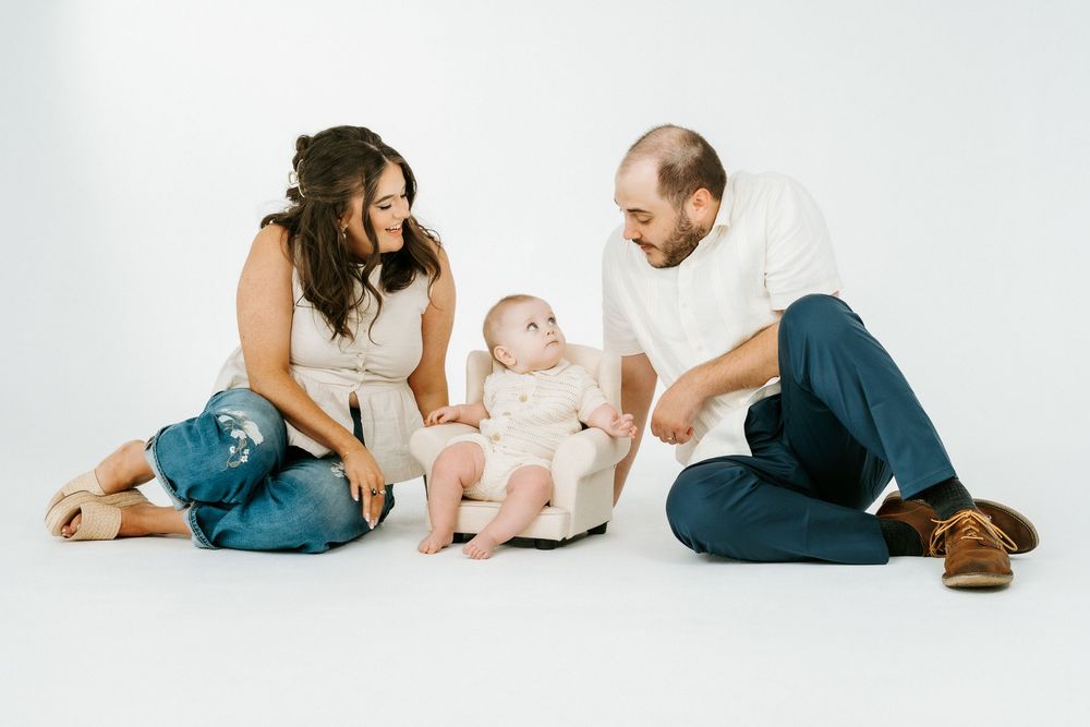 A young family with a baby sitting in a small chair between smiling parents on a white studio background.