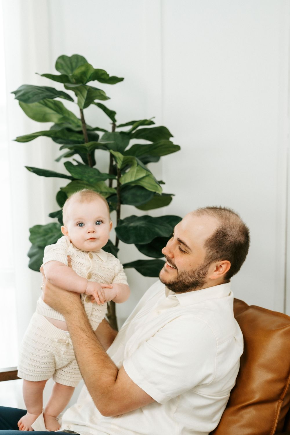 Smiling bearded father holding his baby in a bright white room with a fiddle leaf fig plant in the background.
