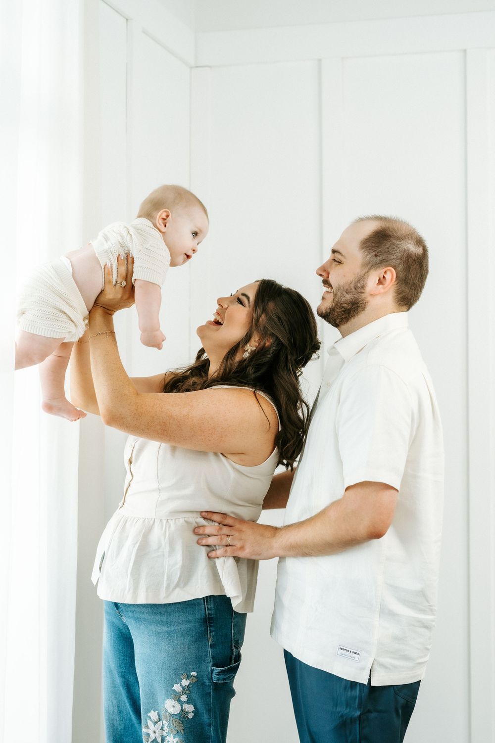 Happy parents lifting their smiling baby in a bright white studio during a professional family photo session.