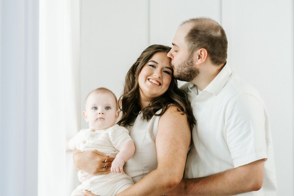 A happy young family with a baby, parents dressed in white, sharing a tender moment in a bright studio setting.