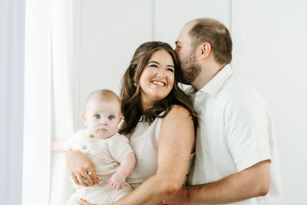 A smiling family with a mother holding a baby while a father kisses her cheek in a bright white studio.