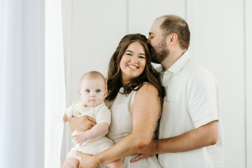 A happy young family with a smiling mother holding a baby while a man kisses her cheek in a bright white studio.
