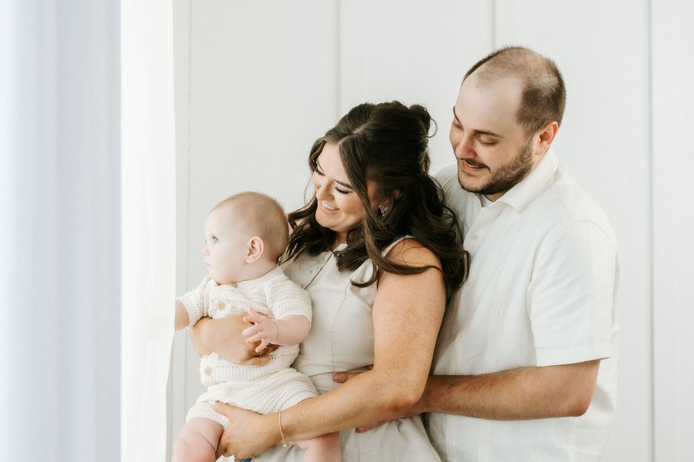 Happy young family with parents holding their smiling baby in a bright, airy white studio setting.