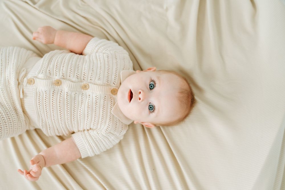 Adorable baby in cream knit romper lying on white bedding, looking up with bright blue eyes.