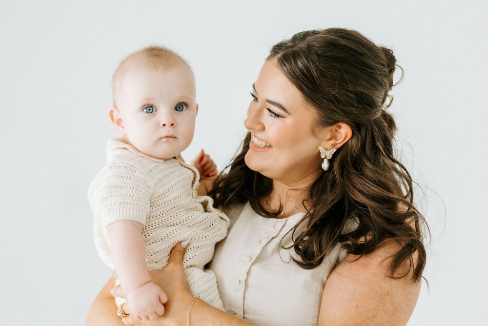 A smiling dark-haired woman holds an adorable baby in a cream knit outfit against a white background.
